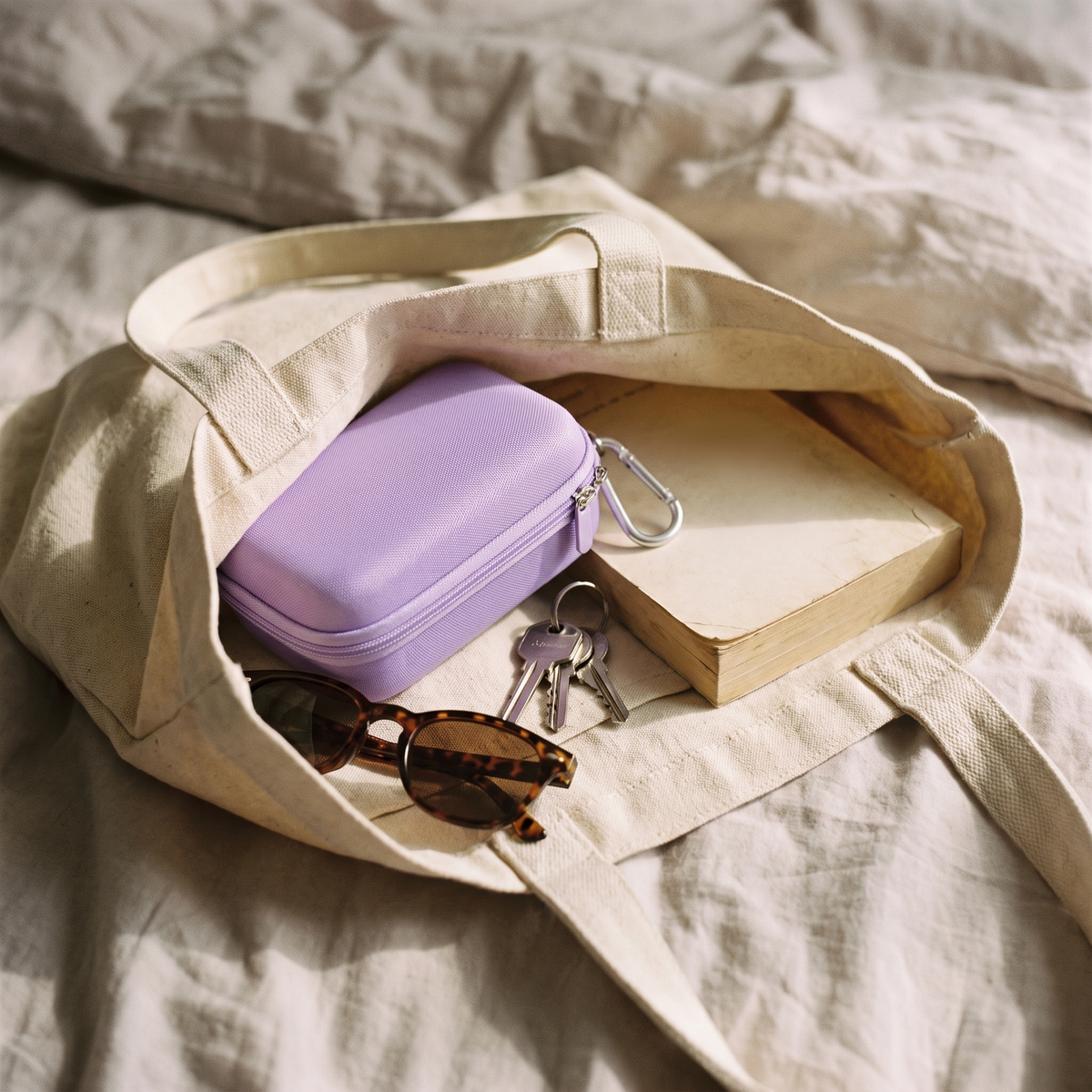 Purple hard shell camera case, zipped, inside a canvas tote bag with keys, sunglasses and a book